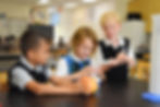A link to the Saint Nicholas Catholic School Elementary School Academics Page. The image shows three elementary school students, two boys and a girl, working on a chemisty experiment.