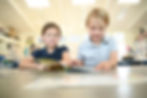 This image links to the Saint Nicholas Catholic School Admissions page. The photo depicts two elementary school students sitting at their desks and reading a book together at Saint Nicholas School  in Los Altos California