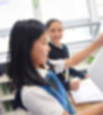 A link to the Academics Page for Saint Nicholas Catholic School. The photo depicts two elementary school girls. One stdent holds up her writing, the other is poised to write more as she looks on.
