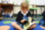 A link to the Saint Nicholas Catholic School Academics page. The image shows a girl in kindergarten  flipping through a book during reading time while sitting comfortably on the floor.