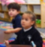 A link to the Saint Nicholas Catholic School Transitional Kindergarten and Kindergarten Acacemics page. The image shows a girl and a boy in TK holding markers and listening intently.
